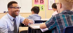 Image of teacher smiling, talking to a student at his desk about accessible instructional materials.