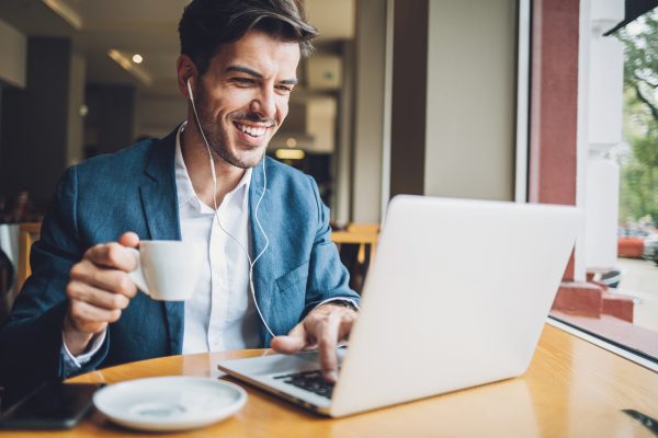 An image of a man in a coffee shop. He’s smiling, listening to audio through earbuds and typing on his laptop.